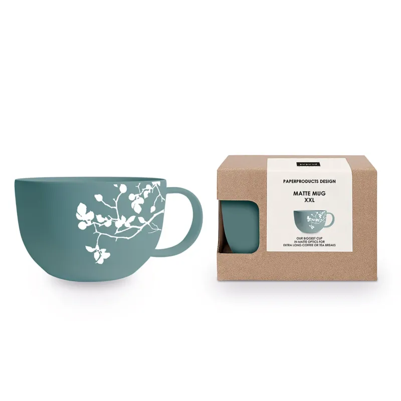 Eine eucalyptusfarbene XXL-Tasse mit weißem Zweigmotiv steht neben ihrer braunen Verpackung.